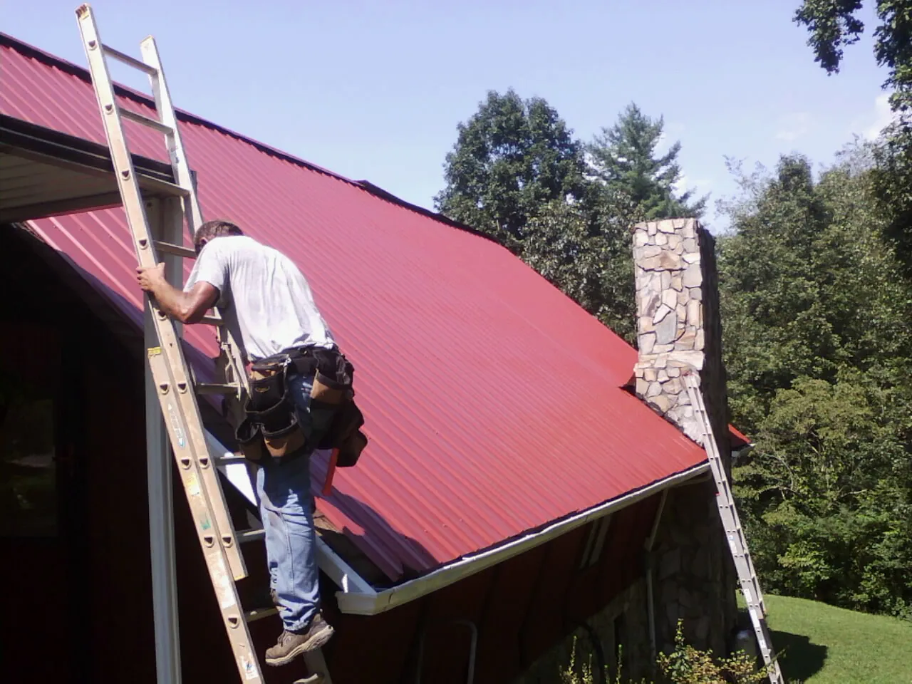 Roofer on a ladder beside a red metal roof and stone chimney — Soffit & Fascia Repair by Roofing Brothers in Idleyld Park